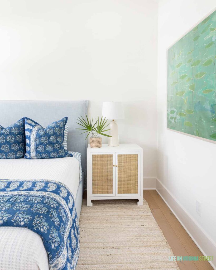 A coastal bedroom with a white jute rug, light blue upholstered bed, blue summer bedroom decor, a white and rattan nightstand, and a cane wrapped vase filled with saw palm fronds.