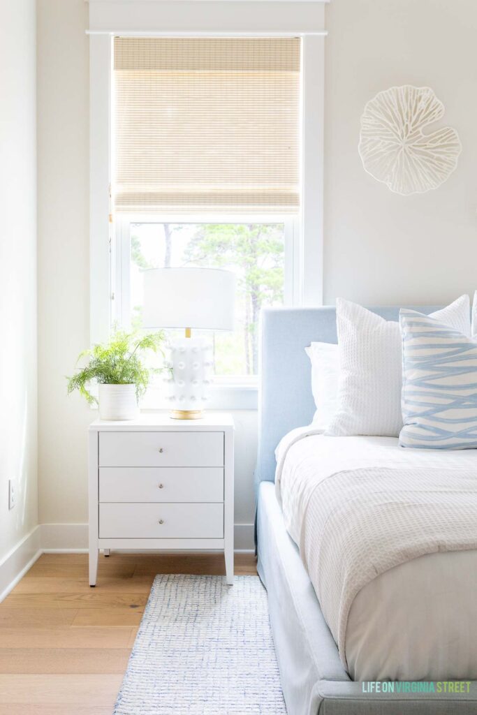 Coastal bedroom ideas for creating a serene space you love! This coastal bedroom includes a light blue linen bed, white nighstands, light blue patterned rug, coral wall art, white designer look for less, woven roman shades, and walls painted Sherwin Williams White Duck.