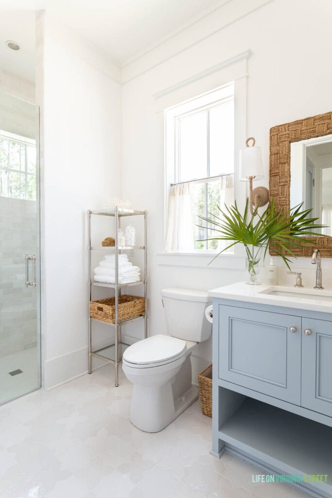 A coastal bathroom featuring blue gray cabinets, woven mirror, woven sconces, cafe curtains, coastal accessories, a walk-in shower, and star and cross floor tile.