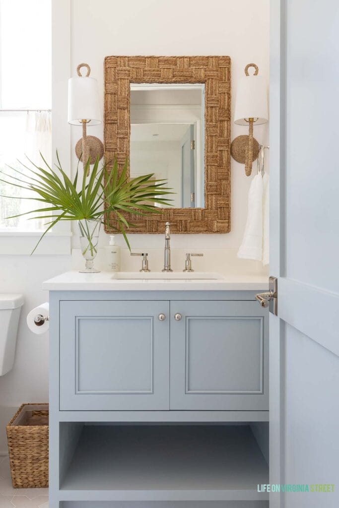 A coastal bathroom featuring blue gray cabinets, woven mirror, woven sconces, coastal accessories, and polished nickel hardware.