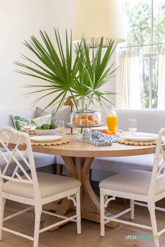 A coastal dining nook featuring a blue and white striped banquette, light wood dining table, white chippendale dining chairs, and styled with palm fronds.