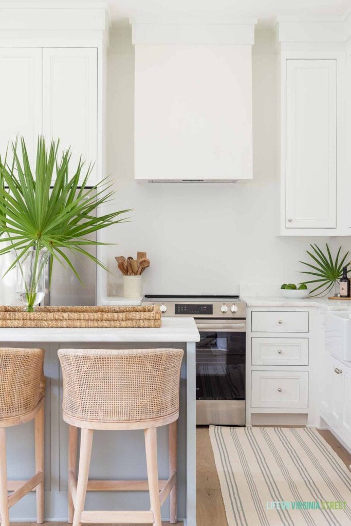 A coastal carriage house kitchen with a white Venetian plaster range hood, white permiter cabinets, a kitchen island painted Benjamin Moore Silver Gray, a blue striped runner rug, and coastal decorative accents.