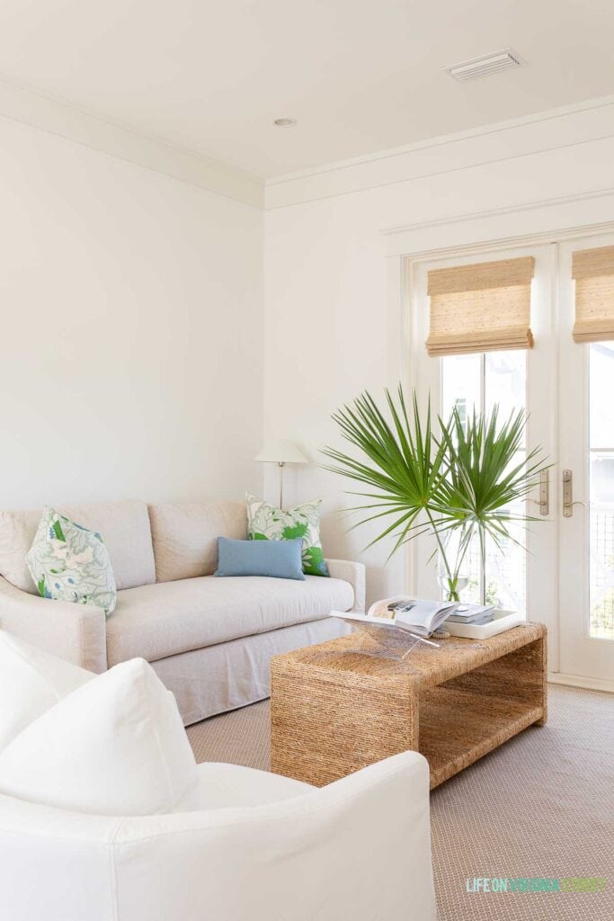 A coastal carriage house living room featuring a linen sleeper sofa, white barrel swivel chairs, a seagrass coffee table, woven roman shades, and coastal accents.