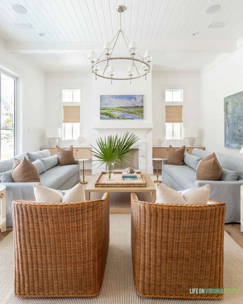 A classic coastal living room with Benjamin Moore Chantilly Lace painted walls, light blue linen sofas, woven swivel chairs, a ring chandelier, a light wood coffee table and decorated with fan palms and white coral.