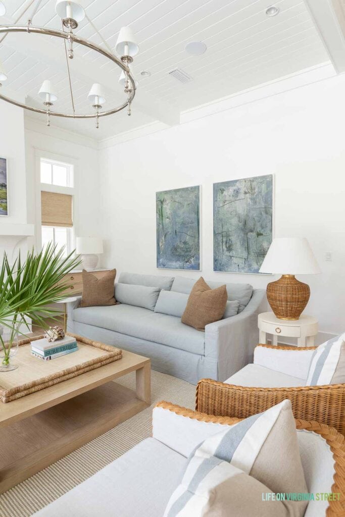 A classic coastal living room with Benjamin Moore Chantilly Lace painted walls, oversized blue and green abstract art, light blue linen sofas, woven swivel chairs, a ring chandelier, a light wood coffee table and decorated with fan palms and white coral.