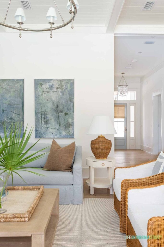 A classic coastal living room with Benjamin Moore Chantilly Lace painted walls, light blue linen sofas, woven swivel chairs, a ring chandelier, a light wood coffee table and decorated with fan palms and white coral. The front door in the distance is painted Benjamin Moore Silver Gray.