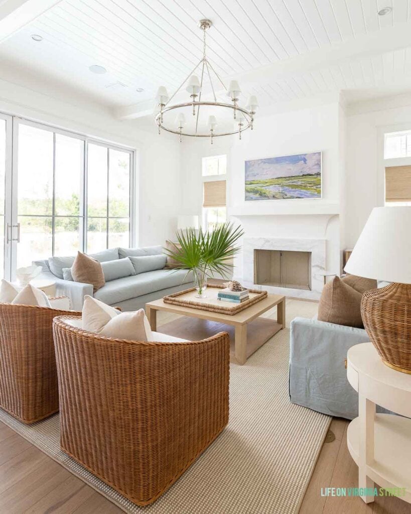 A classic coastal living room featuring large glass sliding doors, with Benjamin Moore Chantilly Lace painted walls, light blue linen sofas, woven swivel chairs, a ring chandelier, a light wood coffee table and decorated with fan palms and white coral.