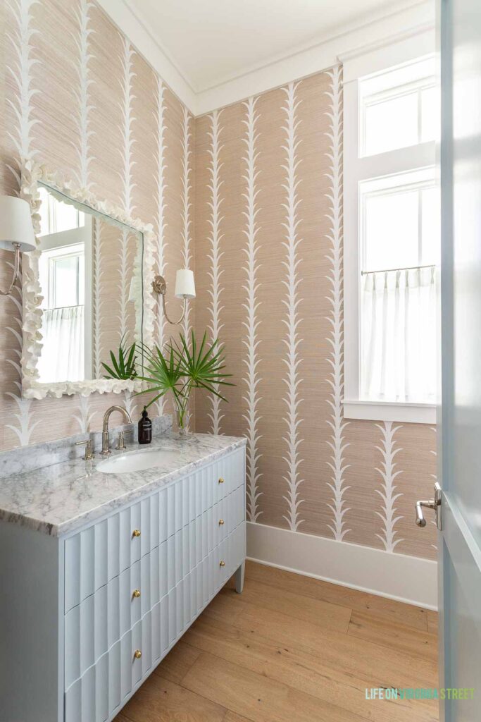 A classic coastal powder bathroom featuring a light blue fluted vanity, natural sisal wallpaper, coral mirror, polished nickel sconces, white cafe curtains, and white oak hardwood floors.