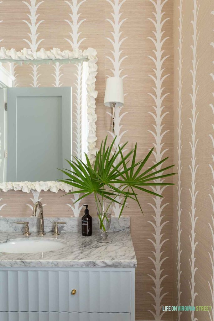 A classic coastal powder bathroom featuring a light blue fluted vanity, natural sisal wallpaper, coral mirror, polished nickel sconces, and fresh fan palms in a glass vase.